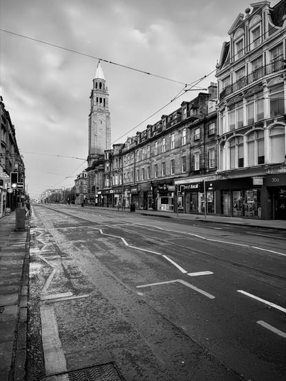 The image shows a street scene captured in black and white. It features a broad street lined with tram tracks, and a mix of historic and classic architecture. Dominating the skyline is a tall tower, likely part of a significant building, possibly a church or municipal structure, typical of European architecture. The street is relatively empty, giving the scene a slightly deserted look. There are shops on the ground floors of the buildings, including what appears to be a local business named "Bayne's". The overall atmosphere is calm, possibly suggesting early morning or a non-peak hour. The connected overhead tram lines suggest public transit integration in this urban setting.