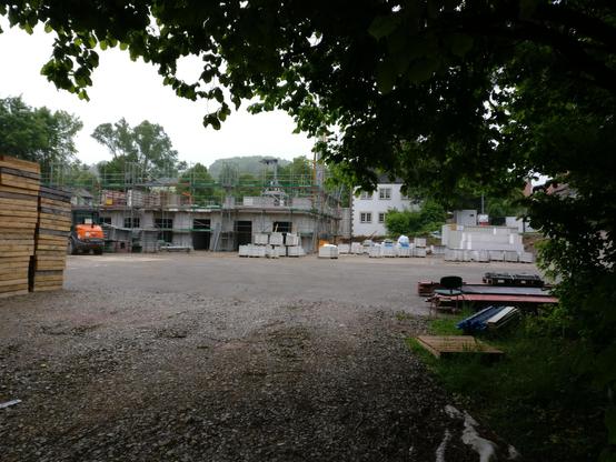 Bauplatz eines institutionellen Neubaus. Die untere Bildhälfte ist dominiert von einem Schotterplatz, am gesamten oberen Bildrand erstreckt sich von rechts her das Blattgrün eines Baums. Ganz links sind zwei Stapel leerer Holzpaletten zu sehen. Hinter dem Schotter wächst links der Neubau, rechts ist - teils verdeckt vom Baum - ein bestehendes, weißes Gebäude zu sehen. Ziemlich nah liegen rechts wenige Baumaterialien, davor steht ein einzelner, grüner Stuhl mit Armlehnen.