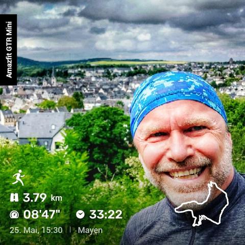 Eine Person mit blauem Stirnband und grauem Oberteil steht vor einer grünen Landschaft mit Bäumen und Sträuchern. Im Hintergrund erstreckt sich eine Stadt mit vielen Häusern, eingebettet in eine hügelige Umgebung. Der Himmel ist bewölkt. Auf dem Bild sind Informationen zu einer Laufaktivität zu sehen: 3,79 km Distanz, eine durchschnittliche Pace von 08:47 Minuten pro Kilometer und eine Gesamtzeit von 33:22 Minuten. Die Aktivität fand am 25. Mai um 15:30 Uhr in Mayen statt. Oben links ist der Schriftzug 'Amazfit GTR Mini' zu sehen