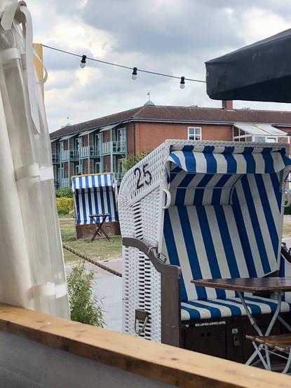 Win Strandkorb mit der Zahl 25 auf der Seite im Vordergrund mit Klapptisch davor. Etwas entfernt im Hintergrund steht ein weiteres Exemplar, auch mit blau-weißem Bezug innen, weiß außen. Ein Appartement-Haus mit Balkonen dahinter.