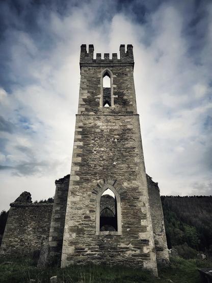 A low-angle shot of the tall, weathered stone tower of an old ruined church against a dramatic, cloudy sky. The tower has several empty arched window openings, and parts of the surrounding ruined walls are visible. To the right, a dark line of trees can be seen.