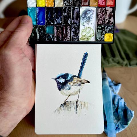 Watercolor sketch of a fairy wren, painted in vivid blues and browns, perched on a stump; artist's hand holds a compact watercolor palette above.