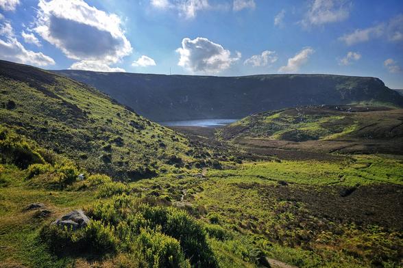 A wide shot of a valley with lush green and brown hills leading down to a lake under a partly cloudy blue sky.