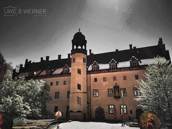 Historic view of the Lutherhaus in Wittenberg, Germany, with its central tower and gabled roof under a dramatic sky. Trees frame the courtyard, where benches and round boards with religious imagery hint at a cultural or historical event.