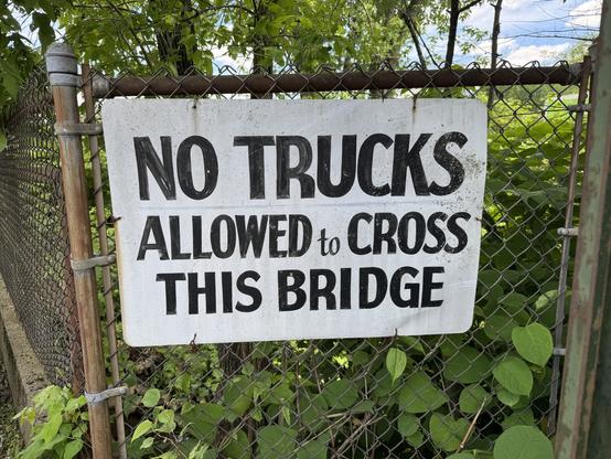 A weathered sign on a chain-link fence reads, "NO TRUCKS ALLOWED to CROSS THIS BRIDGE," surrounded by greenery and trees.