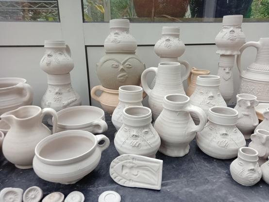 A collection of unfired pottery, like bowls, cups, a dish and many decorated jugs with sculpted faces.