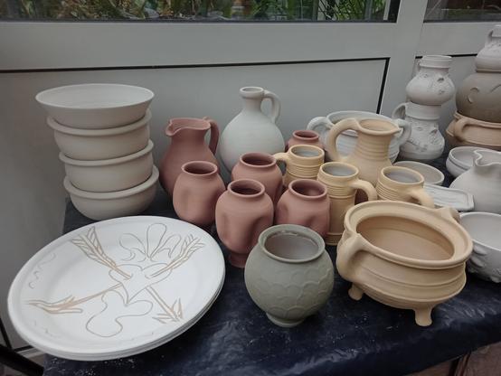 A collection of unfired pottery, like bowls, cups, a dish and many decorated jugs with sculpted faces.