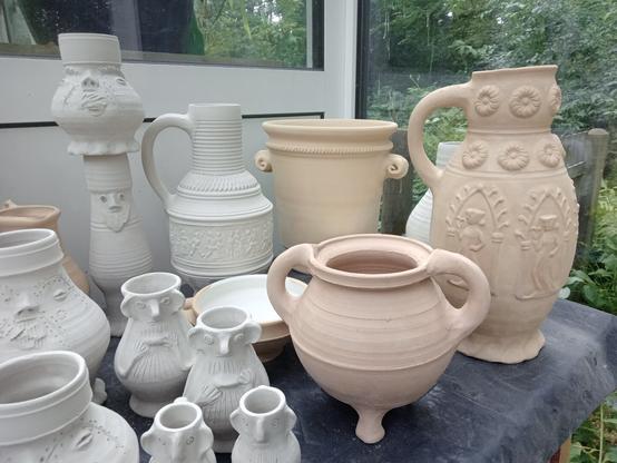 A collection of unfired pottery, like bowls, cups, a dish and many decorated jugs with sculpted faces.