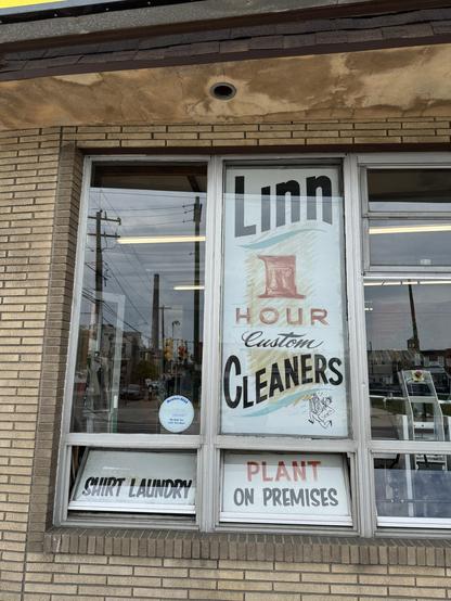 Window display of a cleaners featuring the name "Linn" and the phrase "1 Hour Custom Cleaners." Additional signs indicate "Shirt Laundry" and "Plant on Premises." The building has a brick exterior.