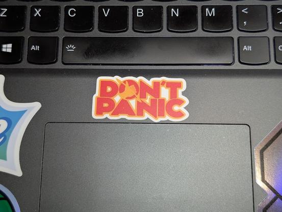 Photo of a sticker on a laptop case below the keyboard, it reads "Don't Panic" with the hole in the "o" containing a hand with the fingers closed and thumb raised, the common hitchhiking gesture. The "o" also has a ring around it like a planet.