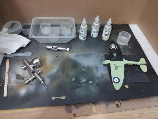Maqueta de un Spitfire MkI  boca abajo recien barnizada. A la izquierda hay un aerógrafo desmontado para limpiarlo, y tras la maqueta hay tres botes de 20ml: el barniz, disolvente para pinturas acrilicas y un mejorador de flujo para aerografia.