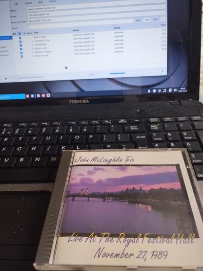 A 1989 CD of the John McLaughlin Trio live at the Festival Hall London sits on an old Toshiba laptop with the lid open showing the Rhythmbox app in the process of ripping the tracks to mp3.