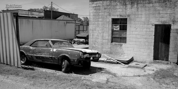 A beat up old 1967 Oldsmobile Cutlass Supreme sits behind a concrete block window next to an alley.