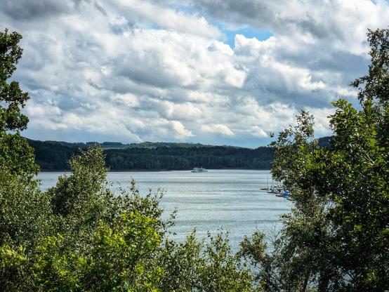 Das Bild zeigt eine idyllische Landschaft mit einem ruhigen See, der von dichtem Grün umgeben ist. Im Vordergrund sind dichte Bäume zu sehen, die den Blick auf den See schützen. Der See erstreckt sich weit in die Ferne, wo sich eine grüne Hügellandschaft erstreckt. Im Hintergrund ist ein großes Schiff auf dem Wasser zu erkennen, das sich langsam bewegt. Der Himmel ist bedeckt mit wechselnden Wolkenformationen, die einen dramatischen Kontrast zu dem ruhigen Wasser bilden. Die Farben sind lebendig, mit verschiedenen Grüntönen der Bäume und dem blauen Wasser des Sees. Die Szene vermittelt ein Gefühl von Frieden und natürlicher Schönheit.

[by AltBot]