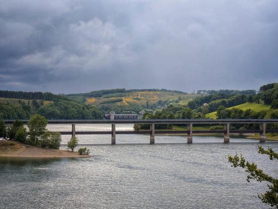 Das Bild zeigt eine ruhige Landschaft mit einem breiten Fluss, der von einem großen, modernen Brückenkranz überquert wird. Der Himmel ist bedeckt mit dunklen Wolken, was auf bevorstehenden Regen hindeutet. Im Hintergrund erstrecken sich grüne Hügel, die mit Bäumen und Sträuchern bedeckt sind. Auf den Hügeln sind auch gelbe Blüten zu sehen, die das Grün der Vegetation durchbrechen. Ein Gebäude, das wie ein Wasserkraftwerk aussieht, ist am Ufer des Flusses zu erkennen. Die Umgebung wirkt friedlich und naturnah, mit einer Mischung aus natürlichen Elementen und menschlichen Strukturen.

[by AltBot]