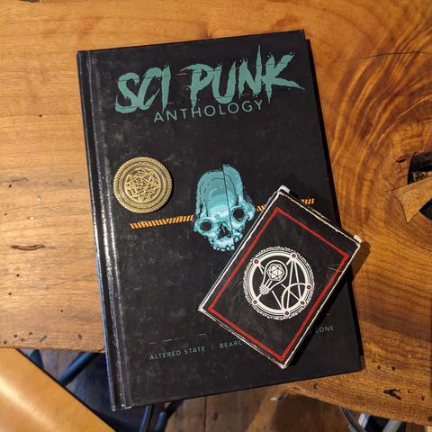 A wooden table with a copy of the Sci Punk Anthology book and the ICRPG Think Deck on top of it