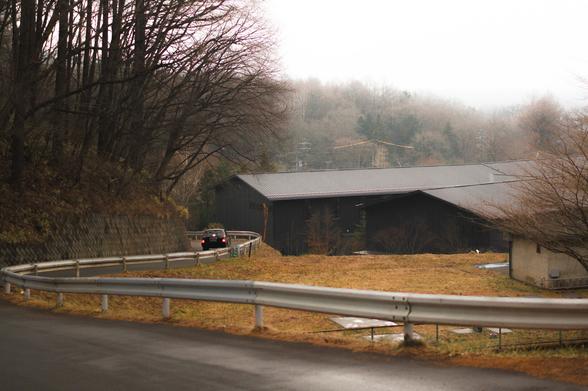 The image features a winding road with a car driving along it, surrounded by trees. There's a field to the right with a large, dark building in the background. The scene has an overcast sky, suggesting a cloudy or foggy day.