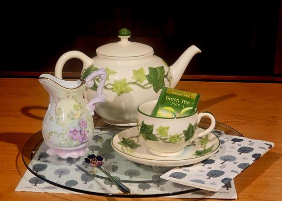 Tea tableau in muted greens on a glass tray with tree-patterned linens and ivy-patterned china, with a separatel cream pitcher lending a hint of pink. The tea is Green tea from Ahmad.