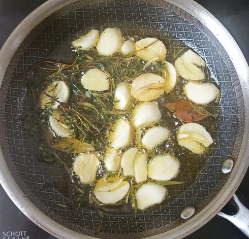 viele Knoblauchzehen + Kräuter in Öl in einer Pfanne
