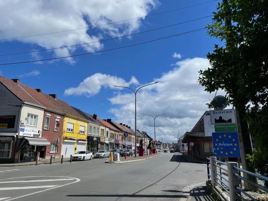 Eine Strasse in einem kleinen Ort, rechts und links Reihenhäuser mit kleinen Läden. Rechts am Strassenrand ein Grenzschild mit der Aufschrift Belgique. Auf der Strasseninsel stehen große Pflanztöpfe mit Bäumen