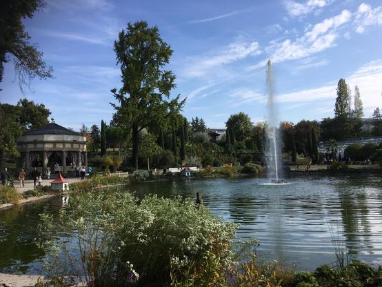 Kleiner künstlicher See mit einer Fontaine. Im Vordergrund Büsche, im Hintergrund blauer Himmel.