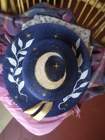 Photo d'un chapeau bleu marine vu du dessus avec un croissant de lune blanc et doré sur le dessus et des feuillages blancs le long du bord.