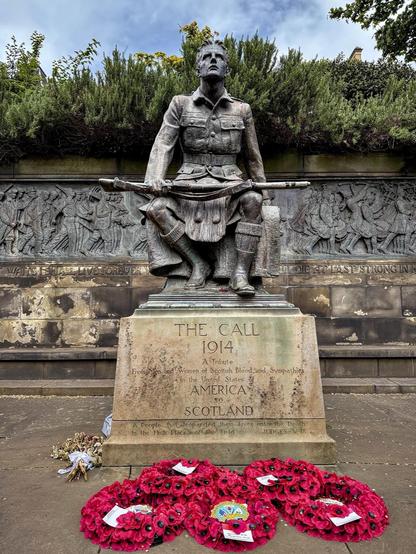 This image shows a memorial statue featuring a kilted soldier standing tall and looking forward, while holding a rifle. The memorial features an inscription that reads "THE CALL 1914" and includes a tribute from the Americans of Scottish blood and sympathies to Scotland. The front of the base cites a line from the biblical Book of Judges: "A People that jeopardized their lives unto the death in the high places of the field."

The statue is adorned with several red poppy wreaths, which are a symbol of remembrance for those who have died in war. The engraving on the base panel below the soldier depicts what appears to be a scene of soldiers in action or marching, suggesting that the memorial could be dedicated to those who served in World War I. The surrounding environment includes greenery and a partly cloudy sky, indicating an outdoor setting, likely within a park or near a public building.