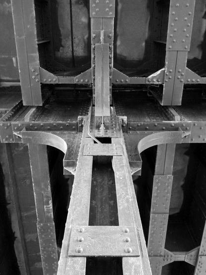 Black and white photo looking up along a steel column at the underside of an elevated railroad line. The column connects to a series of massive steel girders via four large brackets. There are numerous rivets present on each of the structural elements.