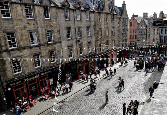 This image depicts a lively street scene in an urban environment that looks historic with old stone buildings and cobblestone paving. The architecture suggests a European city, possibly in the United Kingdom given the style of the buildings and the visible signage. There is festive decoration with numerous colorful bunting flags strung across the street, adding to the vibrant atmosphere.

Various people are seen walking along the street and gathering in small groups, indicating a social or public event setting. To the left side, there's a restaurant named "Mexican Mariachi Restaurant" which contributes a dash of cultural diversity to the scene, marked by its red chairs and exterior décor that stands out against the more subdued colors of the surrounding buildings.

The overall environment appears to be a blend of casual dining, shopping, and possibly tourism, typical of a culturally rich and historical part of a city. The setting is inviting and indicates a section of the city that is pedestrian-friendly and popular among locals and visitors alike.