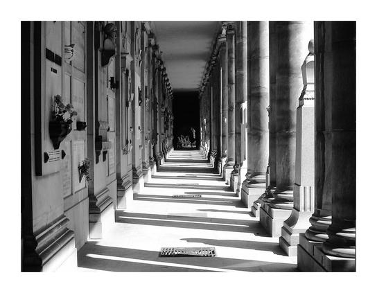 The photograph is a black and white image depicting a long, covered colonnade, similar to a hallway with columns on either side.  The colonnade is quite long, extending into the distance where it appears to darken and become less distinct.  The columns are tall and slender, classical in style, with bases and capitals.  Between the columns, there are walls with what appear to be stone or marble plaques or memorials, possibly tombstones, set into them.  Small floral arrangements are visible in some of these niches.  The floor of the colonnade is paved, and the shadows of the columns are cast long and distinctly on the ground, indicating a strong light source from one side.  A small, rectangular object, possibly a drain cover, is visible in the center of the foreground.  A person is visible in the far distance, adding a sense of scale and depth to the image. The overall impression is one of quiet solemnity and architectural grandeur.