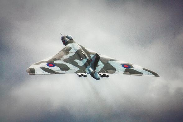 Delta-wing aircraft in flight against a cloudy sky. It has a grey and green camouflage paint scheme and Royal Air Force roundels on both wings. Four jet engines are mounted beneath the fuselage. The aircraft is a British Avro Vulcan bomber, viewed from the rear underside as it ascends.