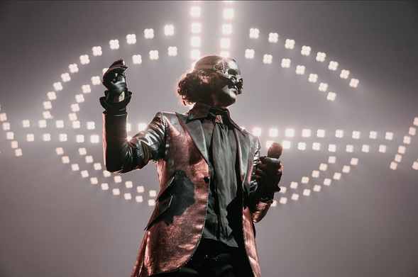 Papa V of the band Ghost is at center. Directly behind and above him is a huge Grucifix (Ghost logo) configured stage lighting fixture.