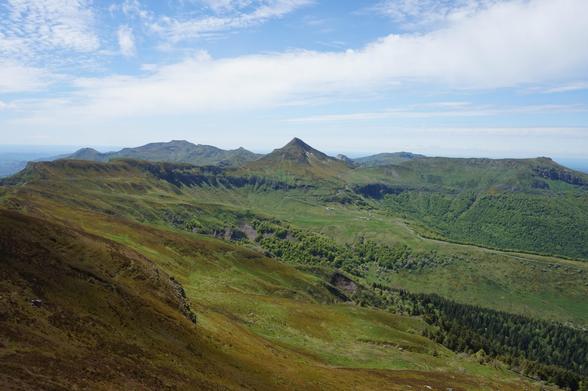 Au premier plan, de vastes prairies d'altitude couvertes de végétation en différentes nuances de vert et de brun. Au loin, des crêtes rocheuses. Le ciel est très nuageux mais les nuages sont clairs.