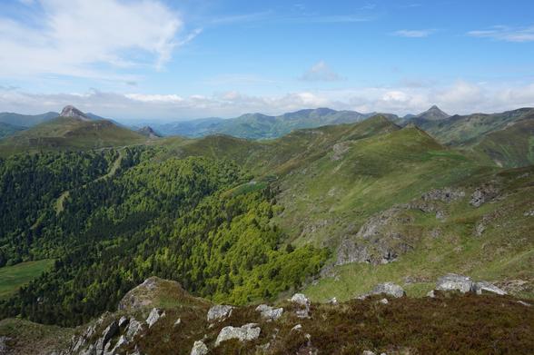Au premier plan, une zone fortement empierrée ; plus loin, plusieurs lignes de crêtes dont les flancs sont verts de végétation. Le ciel est peu nuageux mais des nuages gris sont en approche.