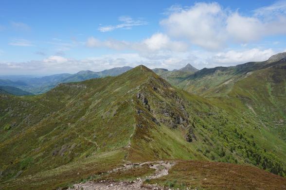 Au premier plan, une zone pierreuse d'où part un sentier menant sur une crête un peu plus loin qui remplit bien le champ de la photo ; on voit encore d'autres reliefs plus loin. Le ciel est un peu nuageux mais les nuages sont clairs.