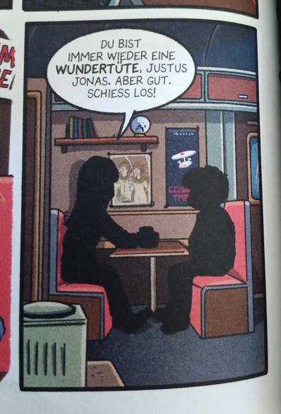 Foto eines einzelnen Comicpanels. Die gleiche Szene wie im ersten Bild, nur von etwas weiter weg. Lys und Justus sind nur als schwarze Umrisse zu sehen. Lys hält ihren Kaffeebecher in der Hand. Das Cosmic Trek Poster ist besser zu erkennen, weil es weniger verdeckt wird.
Lys sagt: "Du bist immer eine Wundertüte, Justus Jonas. Aber gut. Schieß los!"