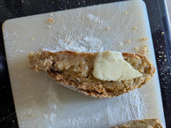The image shows a slice of bread on a white cutting board. The bread appears to be a rustic, whole-grain variety with a rough, textured crust and a dense, grainy interior. A pat of butter is melting on top of the bread, indicating it is freshly cut and ready to be eaten. The cutting board is dusted with flour, and there are crumbs scattered around the slice, suggesting it has been recently cut. The background is a dark surface, possibly a countertop, which contrasts with the light color of the cutting board and the bread. The overall scene suggests a homemade, freshly prepared meal.