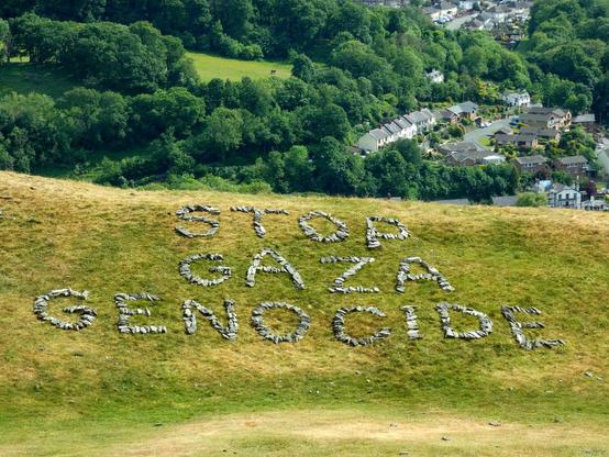The redone message, about 24 x 12 feet in size, "Stop Gaza Genocide", in large lettering made of rocks, on a grassy bank, part of the castle ramparts, above the town and valley far below
