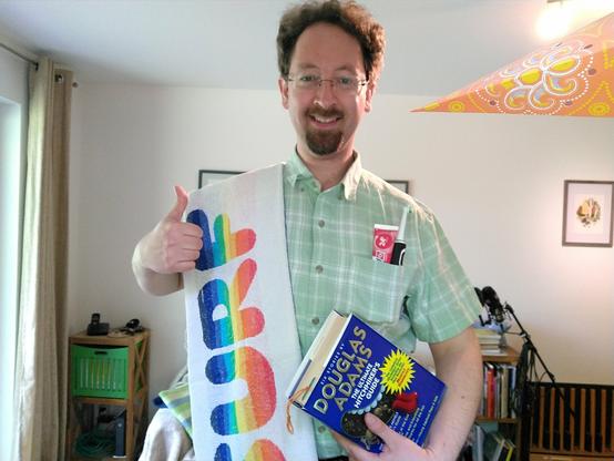 Manuel trägt ein weißes Handtuch über der Schulter, darauf in Regenbogenfarben der Schriftzug „Surf“. In der Hemdtasche eine Tube Zahnpasta und eine elektrische Zahnbürste. Unterm Arm der Sammelband „Douglas Adams – The Ultimate Hitchhiker's Guide“. Der andere Arm zeigt einen Daumen hoch.