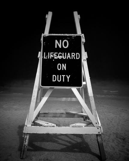 A black and white image showing an empty lifeguard chair with a sign that reads 'No lifeguard on duty.' The chair is on a sandy beach, and the background is obscured by mist with a dark sky above.