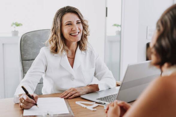Deux personnes attablées face à face : une femme souriante vêtue d'une blouse blanche qui prend des notes en regardant une personne que l'on voit de dos.