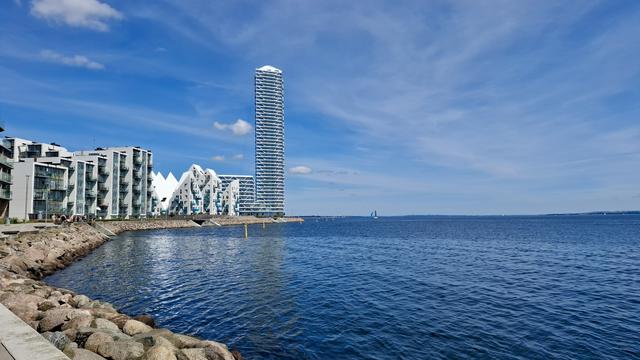 On a pier at the Aarhus Ø docklands area looking out over an expanse of open sea in front of us and to our right. Curving around the edge of the view to our left: the promenade flanked by some of the area’s landmark waterfront apartment blocks on striking angular shapes – the 142-metre high white 'Lighthouse' apartment block; the white-and-blue Isbjerg triangular blocks that are designed like icebergs; rectangular blocks. The sea is dark blue, the summery sky is light blue with white clouds. Edging the promenade, rows of rocks. These are dotted with gradual openings that make it easy to step from the land into the sea. This is the part of the bay where the diving chain and information signs were installed underwater.