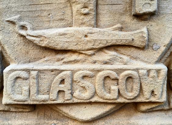 A close up of a section of the City of Glasgow coat of arma featuring the word Glasgow and a dead fish with a ring in its mouth.