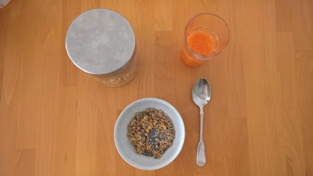 Unten eine gefüllte Müsli-Schalte, rechts daneben ein Löffel, darüber ein mit Saft gefülltes Glas, links davon eine Müslidose, fotografiert senkrecht von oben.