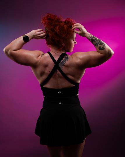 Woman with red hair facing away, wearing a black halter dress. She raises her hands to her hair, displaying muscular arms with tattoos. A tattoo is visible on her upper back. The background is a gradient of pink and purple.