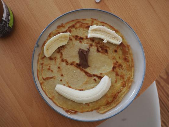 Ein runder Pfannkuchen auf einem runden Teller, der mit Bananen und Nutella als Gesicht verziert wurde.