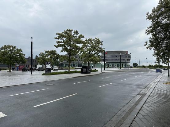 Image of a modern urban street with a wet pavement, featuring a round building and several trees. There are a few parked cars and people near a signboard, under a cloudy sky.