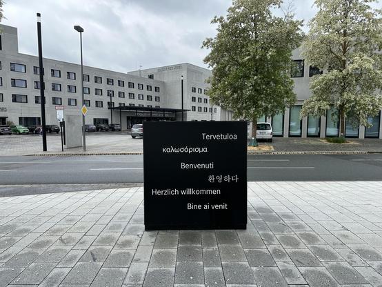 A black welcome sign displaying greetings in multiple languages, including Finnish, Greek, Italian, German, Korean, and Romanian, is positioned in front of a modern hotel building. The hotel features a concrete facade and large windows, with several parked cars visible.