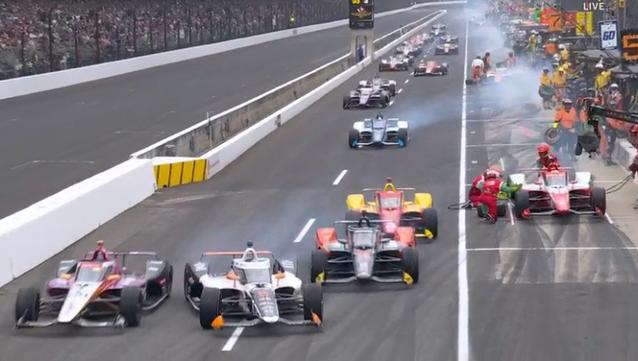 A view down pit lane showing cars, almost three-wide, peeling out of pit boxes and back into the race, with one nearly-avoided collision.