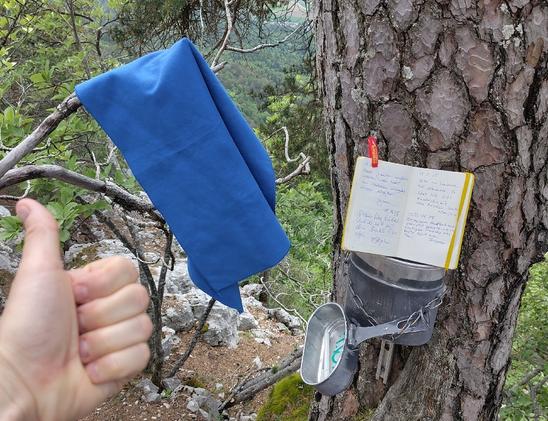 "Gipfel"-Buch n einer Camelle die an einen Baum geschraubt ist. Daneben hängt ein blaues Handtuch am Ast und eine Hand zeigt den Daumen hoch.